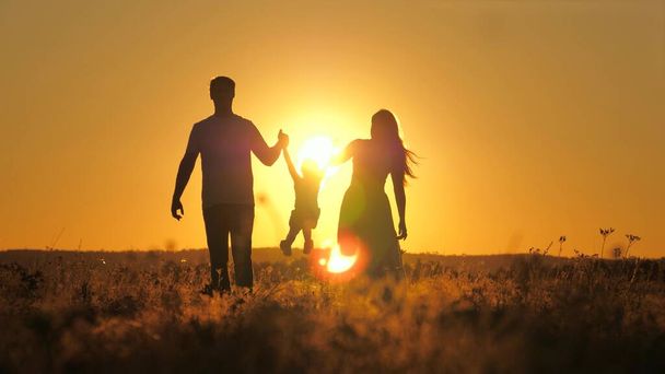 Silhouette d'une famille privée au coucher du soleil dans le ciel, mère, père et enfant marchent à l'extérieur, fille vole dans les bras de papa et maman à l'aube, voyage le soir dans l'éblouissement du soleil dans une belle - Photo, image
