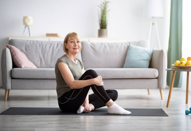 Donna anziana felice che riposa sul tappetino yoga dopo l'allenamento, guardando la fotocamera a casa, spazio libero - Foto, immagini