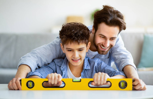 Щасливий батько навчає сина використовувати інструмент рівня для вимірювання поверхні, роблячи ремонт квартири разом
 - Фото, зображення