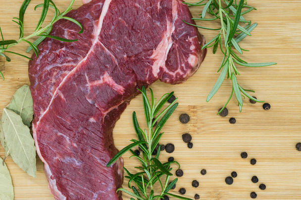 Rindersteak, Blick auf frisches Rindfleisch auf einem Holzbrett mit Rosmarin, schwarzem Pfeffer, Toilettenblatt. - Foto, Bild