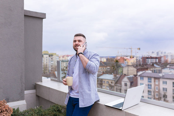 Male businessman, in casual clothes relaxing on the terrace overlooking the city, uses the phone, and uses a laptop while drinking coffee - Photo, Image