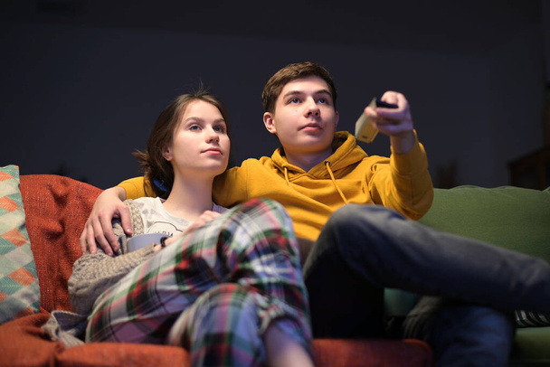 A young guy and his girlfriend sit on the sofa and turn on the TV in the evening. Leisure of a young couple at home in the evening - Foto, Imagen