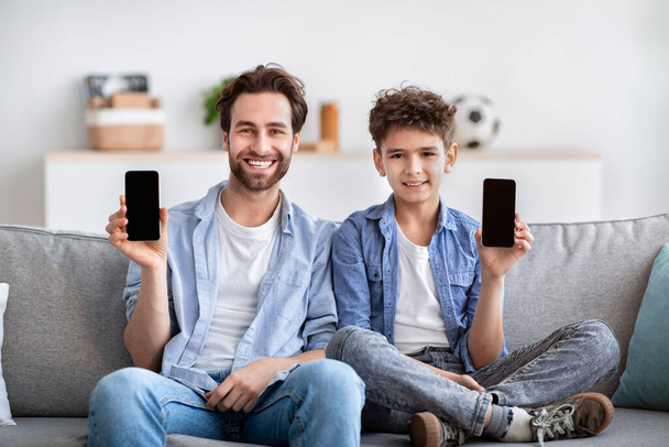 Μας αρέσει αυτή η εφαρμογή. Ευτυχισμένος πατέρας και γιος δείχνουν smartphones τους με λευκές μαύρες οθόνες, κάθεται στον καναπέ στο σπίτι - Φωτογραφία, εικόνα