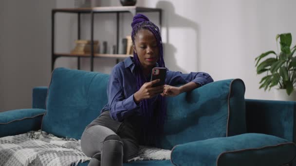 portré fekete tini lány kommunikál video chat okostelefon, hölgy raszta ül a kanapén a modern lakásban - Felvétel, videó
