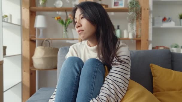 Zamyšlená asijská dáma sedí objímající kolena na pohovce v obývacím pokoji v domě pohled ven s pocitem osamělosti, Smutný depresivní teenager tráví čas sám doma, Společenská vzdálenost, Koronavirus karanténa. - Záběry, video