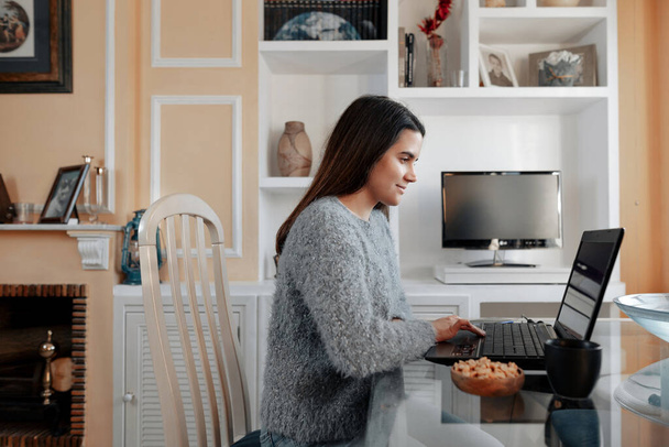 Młoda, pogodna kobieta w swetrze w domu siedzi w salonie i przegląda internet w laptopie. - Zdjęcie, obraz