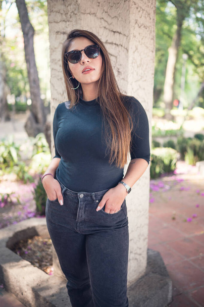 A cool sexy Mexican woman in sunglasses posing with hands in pockets in a garden - Photo, image