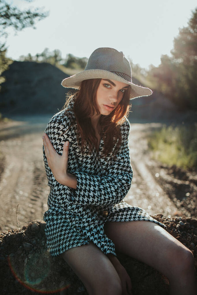 A vertical shot of a young caucasian female in a houndstooth pattern coat and fedora hat sitting on a stone - Fotó, kép
