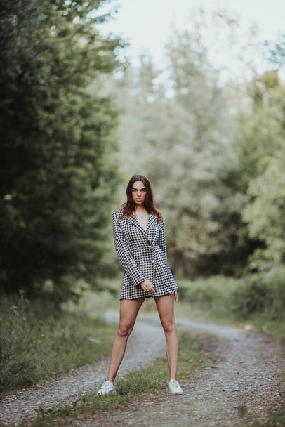 A vertical shot of a caucasian female in a houndstooth pattern dress standing on a forest trail - Foto, afbeelding