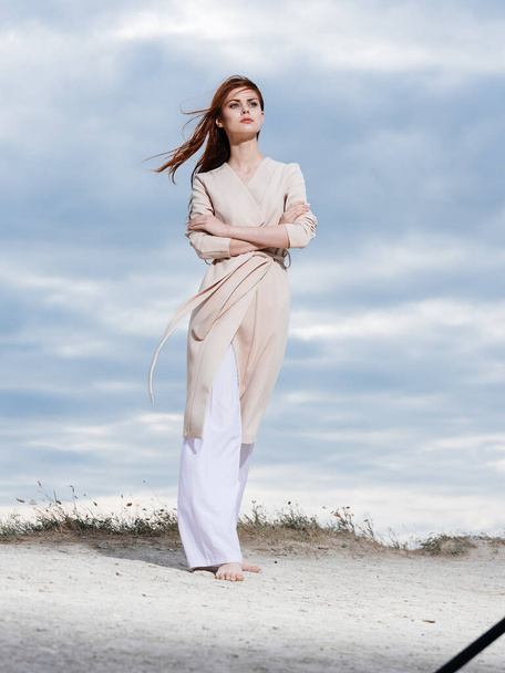 Γυναίκα σε ένα πουλόβερ και σε λευκό παντελόνι στην άμμο μπλε ουρανό σύννεφα φύση. - Φωτογραφία, εικόνα