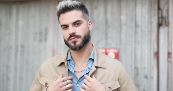 attractive young casual guy in denim shirt fixing beige jacket while confidently looking around and touching beard against metal garage outside - Footage, Video