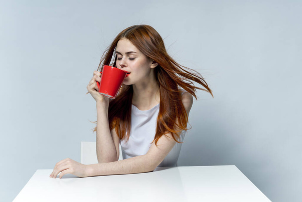 atractiva mujer pelirroja bebiendo de una taza roja mientras está sentada en la mesa - Foto, Imagen