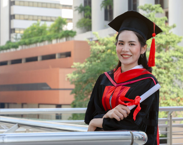 Portrait of happy graduated woman in black graduation gowns holding diploma and standing with arms crossed. Smiling student wearing mortarboard cap looking at camera with building background. - Photo, Image