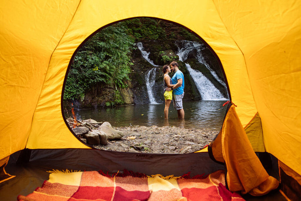 couple standing in the lake waterfall on background view from yellow camping tent. summer activities - Photo, Image
