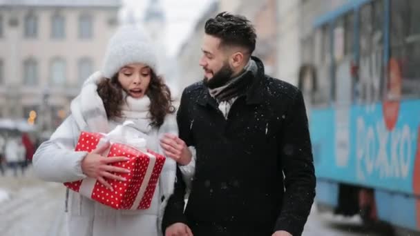 Un uomo e una donna stanno camminando attraverso il centro della città in una nevicata. Una donna sta portando un grande dono. Parlano e ridono. Sullo sfondo passa un tram. 4K - Filmati, video