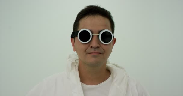 Homme en costume de protection chimique blanc et lunettes rondes noires se tient avec une expression sérieuse. Puis il rit et enlève ses lunettes, au ralenti - Séquence, vidéo