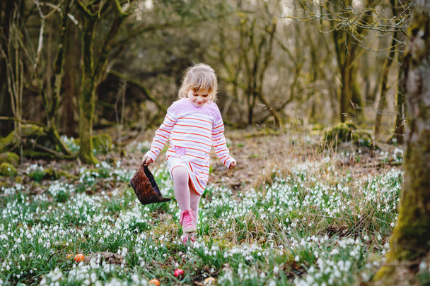 Dziewczynka w różowej sukience polująca na jajka w wiosennym lesie w słoneczny dzień, na świeżym powietrzu. Cute szczęśliwy dzieciak z dużą ilością kwiatów śniegu i kolorowych jaj. Wiosna, chrześcijańskie święto koncepcja. - Zdjęcie, obraz