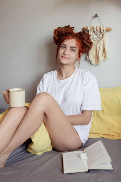 Nagyon csinos, vörös hajú lány, rasztával az ágyon reggel a hálószobában. Egy nőies, gyengéd lány. lány miután reggeli az ágyban tart egy csésze kávét, és könyvet olvas - Fotó, kép