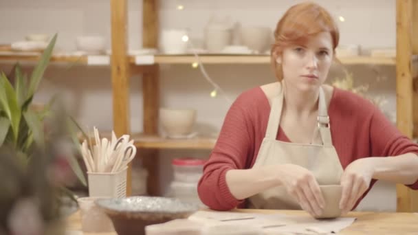 Waist-up portrait shot of young Caucasian woman in apron and casual sweater sitting at table in pottery workshop, sculpting and thinning walls of wet clay bowl and looking at camera with happy smile - Footage, Video