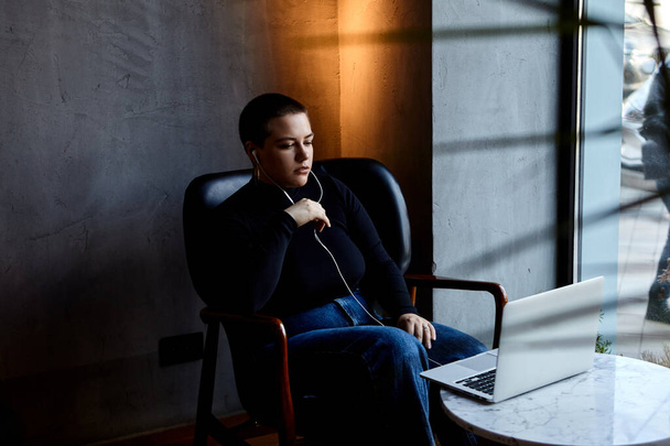 Una chica joven con auriculares, pelo corto y piercing en la nariz está sentada en un café y hablando en un enlace de video por computadora portátil - Foto, Imagen