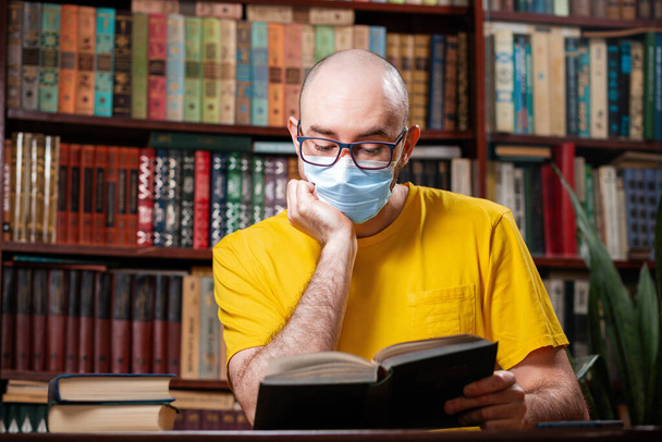 Virus. Un hombre calvo está sentado en gafas y una máscara médica y leyendo un libro. En el fondo hay una biblioteca y estanterías de libros. Concepto de autoaislamiento, aprendizaje a distancia y cuarentena. - Foto, imagen