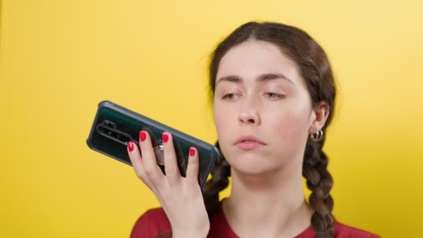 Μια νεαρή όμορφη μελαχρινή γυναίκα μιλάει σε ένα smartphone, ηχογραφώντας ένα φωνητικό μήνυμα στην εφαρμογή. Κίτρινο φόντο. Έννοια επικοινωνίας. - Πλάνα, βίντεο