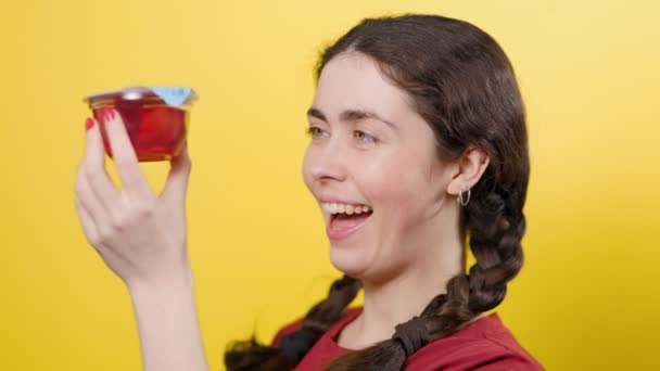 Retrato de una chica bonita abre un frasco de gelatina, huele y quiere probarlo. Fondo amarillo. Postres sin azúcar - Imágenes, Vídeo
