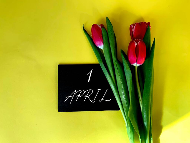 1 april op zwart papier.Daarnaast staan rode tulpen op een gele achtergrond.In de hoek van de foto de schittering van de zon.Kalender voor april. - Foto, afbeelding