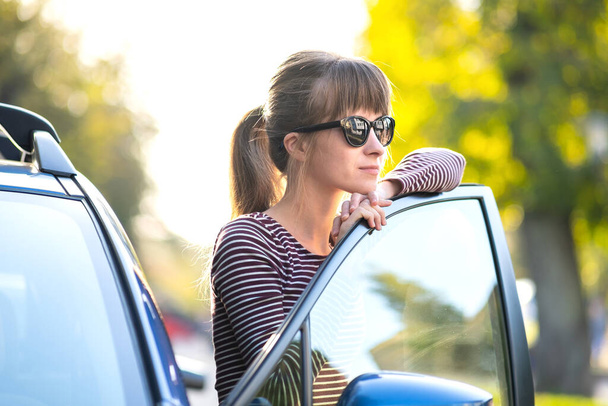 Conductora joven feliz disfrutando de un cálido día de verano de pie junto a su coche en una calle de la ciudad. Concepto de viaje y vacaciones. - Foto, imagen