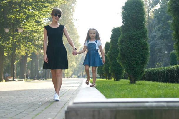 Jeune mère et sa petite fille aux cheveux longs marchant ensemble se tenant la main dans le parc d'été. - Photo, image