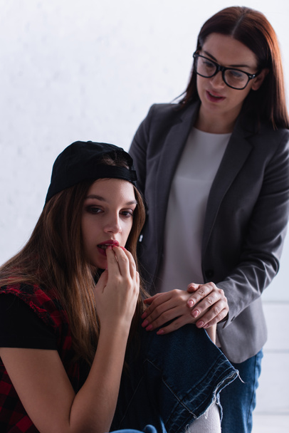 ustaraný dospívající dívka v čepici kousání nehty v blízkosti psycholog na rozmazaném pozadí - Fotografie, Obrázek