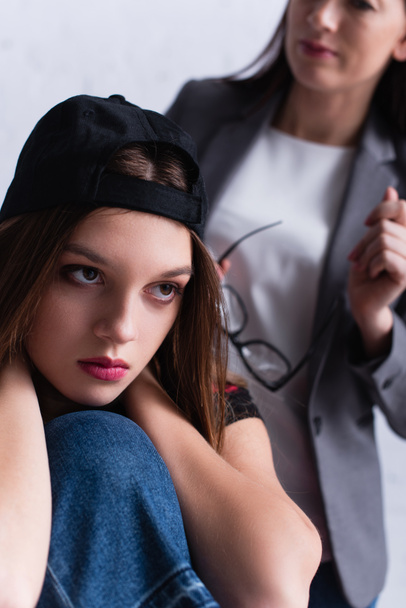 displeased teenage girl in cap near psychologist on blurred background - Photo, Image