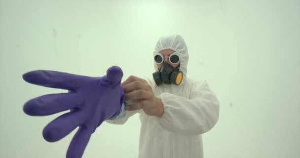 Közelkép az ember fehér vegyi védőruhában és gázálarcban felvesz egy lila színű gumikesztyűt, lassított felvétel - Felvétel, videó