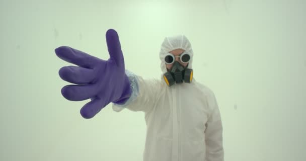 Közelkép az ember fehér vegyi védőruhában és gázálarcban felvesz egy lila színű gumikesztyűt, lassított felvétel - Felvétel, videó
