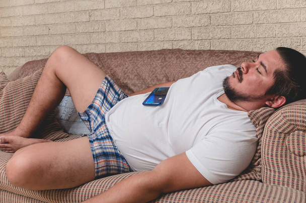 Ein arbeitsloser, arbeitsloser oder unter Quarantäne stehender Mann schläft auf der Couch, sein Handy auf dem Bauch liegend. - Foto, Bild