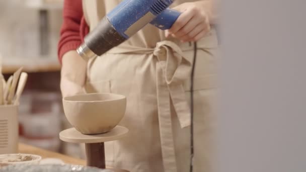 Medium tilting up shot of female pottery artist drying handmade clay bowl with heat gun, putting it down on table, picking up vessel and checking it - Footage, Video