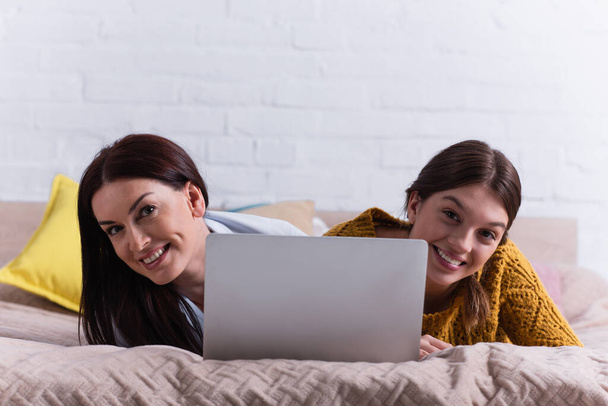 улыбающаяся мать и счастливая девочка-подросток смотрят в камеру рядом с ноутбуком в спальне - Фото, изображение