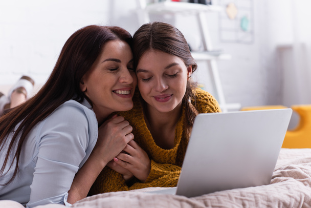 glimlachende moeder en tiener meisje knuffelen in de buurt laptop in slaapkamer - Foto, afbeelding