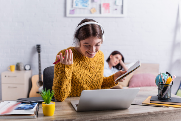 glückliches Teenager-Mädchen in drahtlosen Kopfhörern mit Notizbuch und Stift in der Nähe von Laptop - Foto, Bild