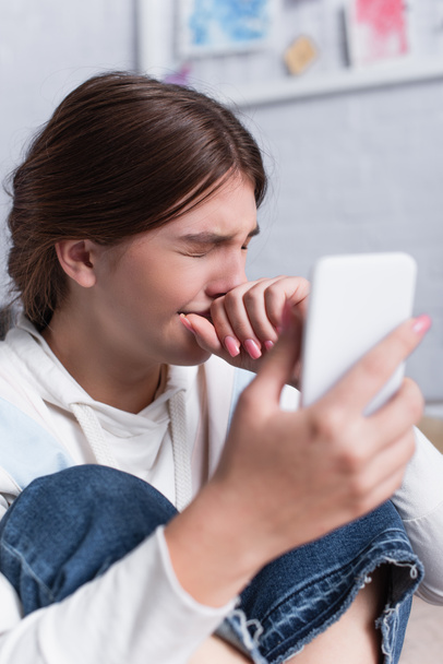 smutna nastolatka dziewczyna obejmujące usta podczas płaczu i trzymając smartfon  - Zdjęcie, obraz