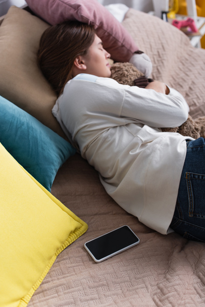 wysoki kąt widzenia smartfona z pustym ekranem na łóżku w pobliżu smutnej nastolatki leżącej z misiem - Zdjęcie, obraz
