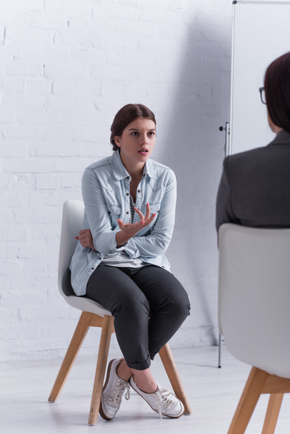 ustaraný dospívající dívka sedí a gestikuluje při pohledu na psycholog na rozmazané popředí  - Fotografie, Obrázek
