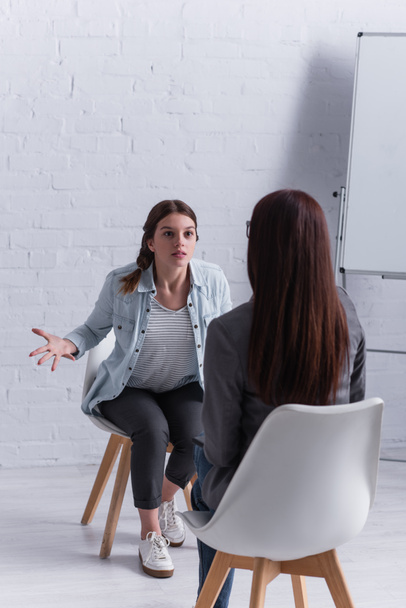 upset teenage girl sitting and gesturing while looking at psychologist on blurred foreground  - Photo, Image