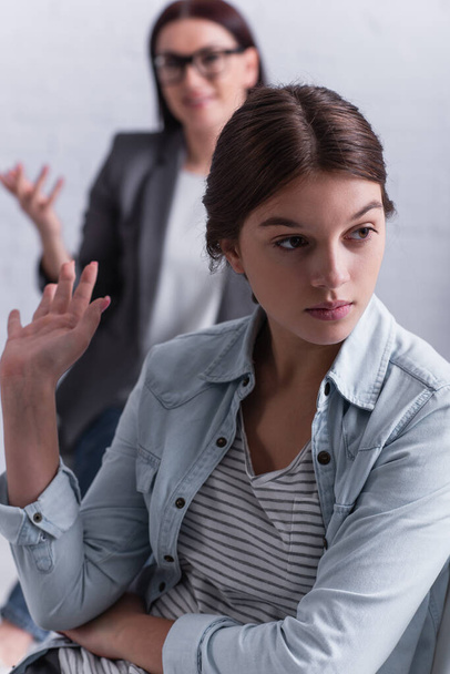 depressed teenage girl looking away and gesturing near psychologist sitting behind on blurred background  - Photo, Image
