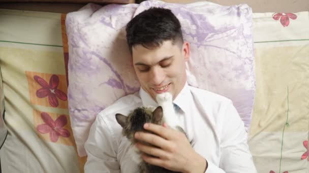 Ofis kıyafetleri içindeki yakışıklı genç adam işten sonra yatağında kedisine uzanıyor. - Video, Çekim