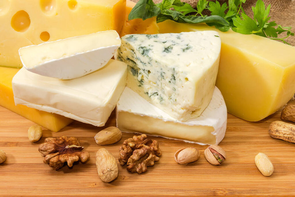 Pieces of various types of soft, semi-hard and blue cheese among the greens and nuts on a wooden surface - Photo, Image