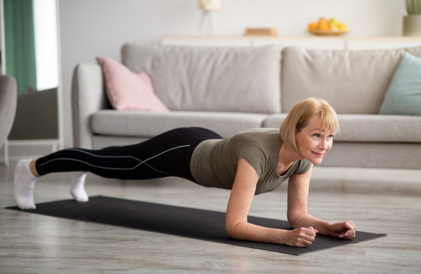 Huiselijk trainingsconcept. atletische senior vrouw staan in elleboog plank op yoga mat thuis - Foto, afbeelding