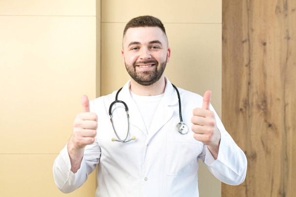 portrait of a friendly doctor in a uniform at the table in the hospital, smiling and looking at the camera. professional physician wearing white medical coat, stethoscope sitting at workplace - Photo, Image