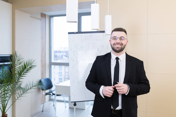 Portrait young bearded man in a formal black business suit with glasses and tie looking at camera. Confident Male a modern office a whiteboard. Manager ceo meeting or conference person, businessman - Fotó, kép