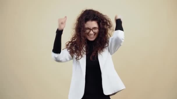 Crazy young woman with curly hair dances with clenched fists waving her hands and head. Young hipster in gray jacket and white shirt, with glasses posing isolated on beige background in the studio. - Footage, Video